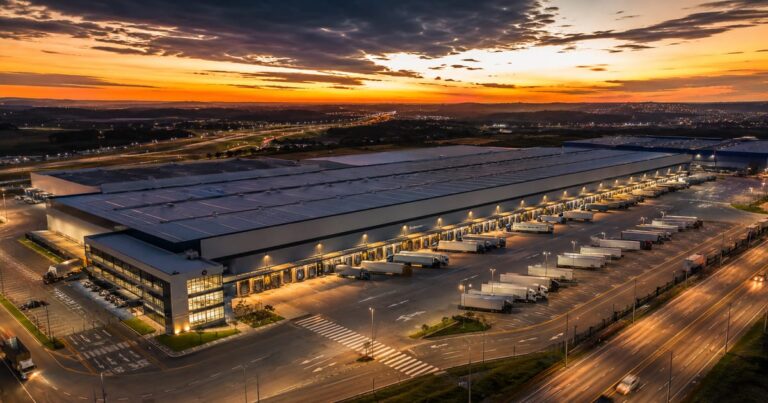Visão aérea de um grande complexo de galpões logísticos modernos com caminhões nas docas ao pôr do sol.